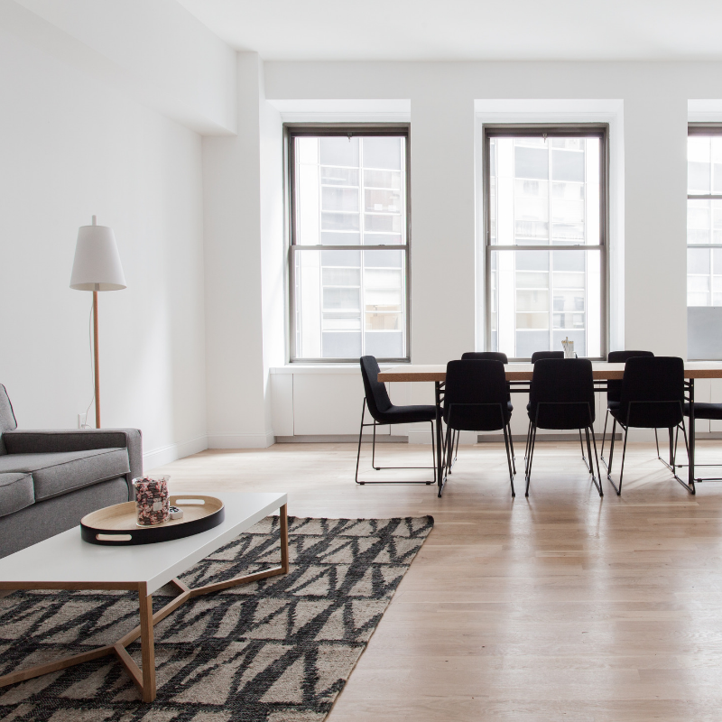 Modern minimalist living room with large windows, hardwood floors, gray sofa, white coffee table, black dining chairs, wooden dining table, patterned area rug, and floor lamp.
