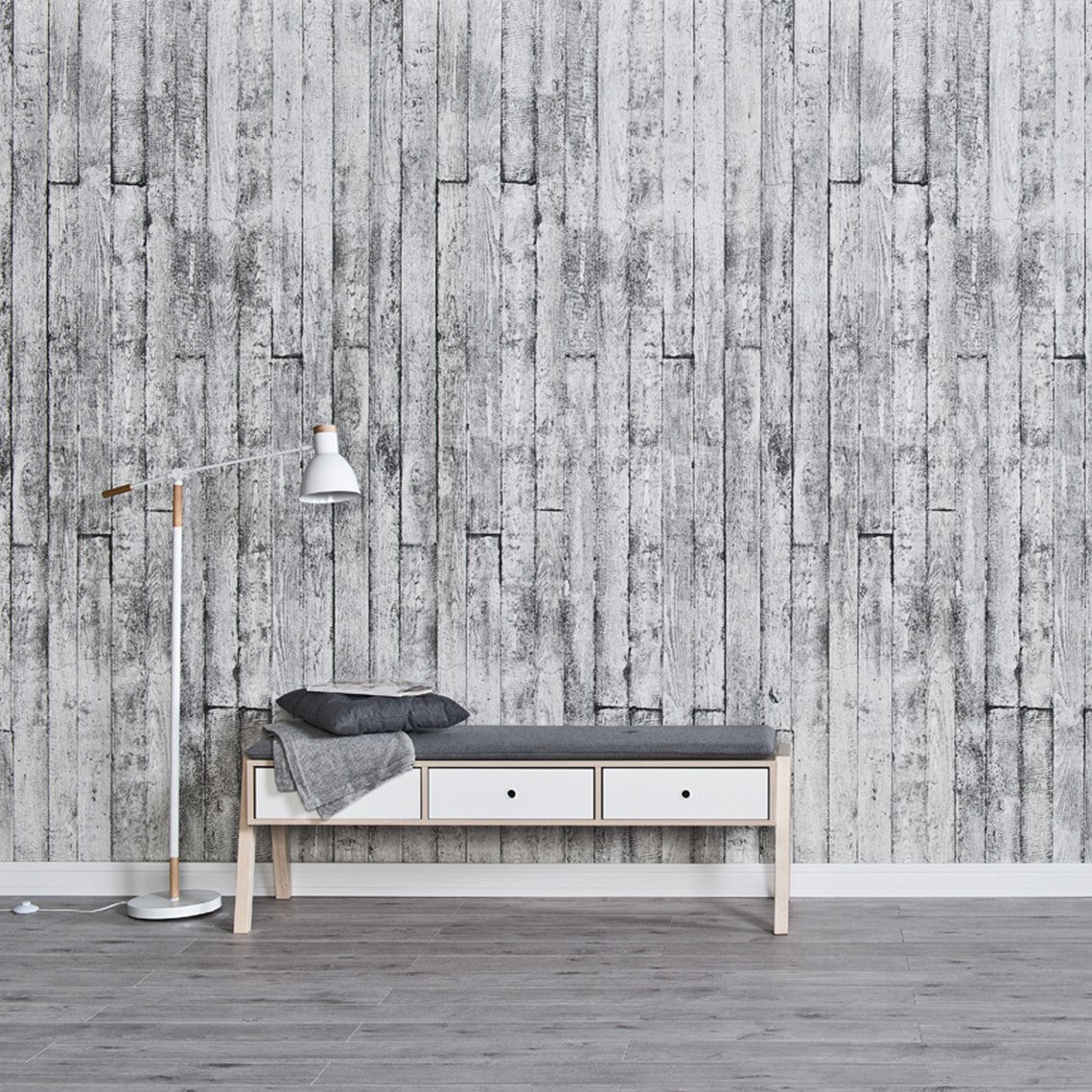 Modern minimalist interior with textured grey wooden plank wallpaper, sleek white and wood bench with storage drawers, cozy gray cushion and throw blanket, stylish floor lamp with white shade and wooden accents, and gray flooring.